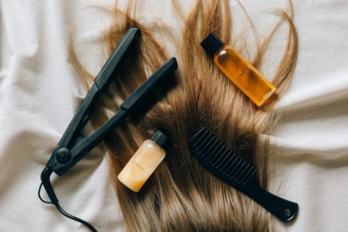 Hair styling tools including straightener, brush, and product laying on top of human hair.