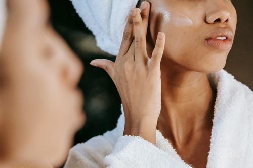 A woman wrapped in a towel applies cream to her face